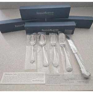 May include: Silverware set from Reed & Barton, including spoons, forks, and a knife, displayed in individual plastic sleeves. The set is presented with dark blue boxes and informational cards. The Reed & Barton logo is visible.