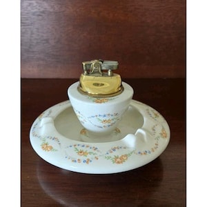 May include: A vintage white ceramic ashtray and lighter set. The ashtray is round with a central lighter holder, both decorated with a floral pattern in orange, blue, and yellow. The lighter has a gold-colored metal top.