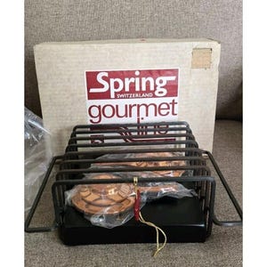 May include: A black metal serving rack with a rectangular base and a wire grid. The rack holds several copper-colored serving dishes, still in their original packaging. The box behind the rack has the words "Spring Switzerland gourmet line".