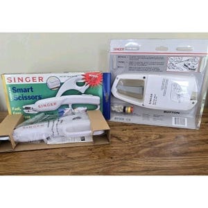 May include: Two Singer sewing tools are displayed: a box of cordless smart scissors and a buttonhole maker. The scissors are white with the text "Smart Scissors" and "Fast, Easy, Free". The buttonhole maker is in a clear plastic package.