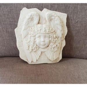 May include: A cream-colored angel wall plaque with a serene face, curly hair, and feathered wings. The angel has a halo and is set against a textured, irregular background. This decorative piece is suitable for home decor.
