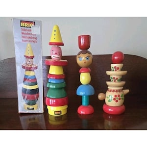 May include: A collection of colorful wooden stacking toys, including a clown, a face, and a teapot design. The toys are made of painted wood and come in various shapes and sizes. The box reads "BRIO" and "Wooden toys".
