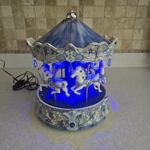 May include: A carousel lamp featuring a blue stained-glass style roof and base with floral patterns. The structure includes white horses on silver poles, illuminated by a bright blue light. Small blue crystal-like pendants hang from the roof edge.