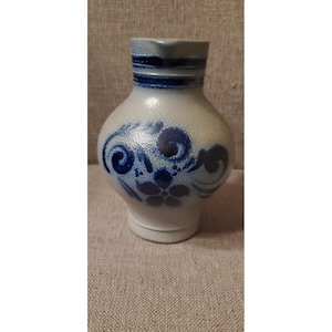 May include: A stoneware pitcher with a bulbous body and a spout. The pitcher is light gray with dark blue floral designs and bands around the neck. The base is white. A decorative piece for the kitchen or dining room.