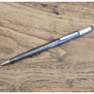 May include: A black pen with a silver tip and a silver end cap. The pen has a textured pattern and the text "U.S. GOVERNMENT" is printed on the side. The pen is lying on a wooden surface.