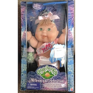 May include: A Cabbage Patch Kids Millennium Celebration doll in its original packaging. The doll has blonde hair, green eyes, and a pink bow. It wears a sash that says "2000". The packaging includes a baby bottle and the words "Millennium Celebration".