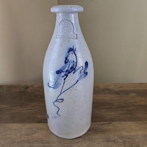 May include: A light gray stoneware bottle with a speckled texture and a narrow neck. The front features a blue hand-painted abstract design and an embossed stamp that reads HANDMADE ROWE POTTERY WORKS CAMBRIDGE WIS.