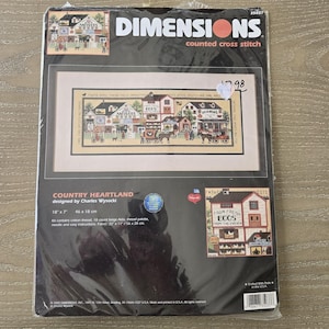 May include: A Dimensions counted cross stitch kit titled "Country Heartland" designed by Charles Wysocki. The kit includes a detailed cross-stitch pattern of a quaint village scene, measuring 18 x 7 inches.