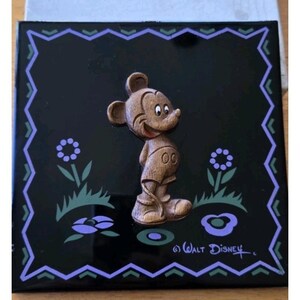 May include: A square, black decorative tile featuring a 3D relief of Mickey Mouse. The tile has a border with a purple and green design, with floral accents. The Walt Disney signature is in the bottom right corner.