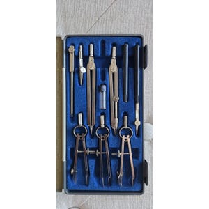 May include: A set of silver drafting tools, including compasses, dividers, and pens, neatly arranged in a blue velvet-lined case. The tools are made of metal and are designed for precision drawing and measurement. The case is rectangular with a black exterior.