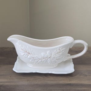 May include: A cream-colored ceramic gravy boat with an attached rectangular base. The exterior features a raised, embossed pattern of oak leaves and acorns. It has a curved handle on one side and a wide pouring spout on the opposite end.