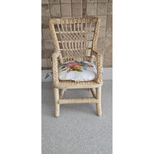 May include: A small, light-colored wicker chair with a high back and armrests. The chair features a woven design and a square cushion with a floral pattern in pink, yellow, and blue. The chair is suitable for a doll or small child.