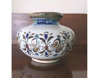 Könnte beinhalten: Eine Keramikvase mit bauchigem Körper und kurzem Hals. Die Vase ist weiß mit einem floralen Muster in Blau, Braun und Grün. Der Rand ist braun und blau. Die Vase steht auf einer dunklen Holzoberfläche.