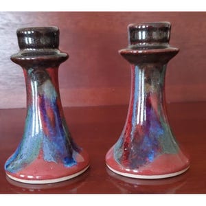 May include: Pair of ceramic candle holders with a flared base and a dark brown top. The candle holders have a colorful glaze with red, blue, and green hues. The base is red, and the overall design is artistic and decorative.
