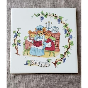 May include: Square ceramic tile featuring a charming illustration of three mice gathered around a fireplace. One mouse reads a book labeled "Story Time" to the other two. The tile is framed by grape vines and the words "Story time".