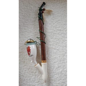 May include: A decorative tobacco pipe with a white ceramic bowl featuring a red floral design and the word "Austria". The pipe has a dark wooden stem wrapped with green twine, a black mouthpiece, and a small feather accent.