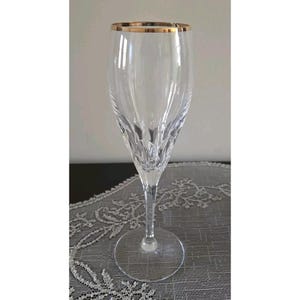 May include: A clear champagne flute with a gold rim. The glass has a long stem and a wide, round base. The body of the glass has a textured, vertical design. The glass is sitting on a gray, patterned surface.