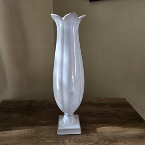 May include: A tall, white ceramic vase with a glossy finish, featuring a flared, petal-like rim and a slender, vertically ribbed body that tapers down to a pedestal base. The vase stands upright with a smooth, reflective surface throughout.