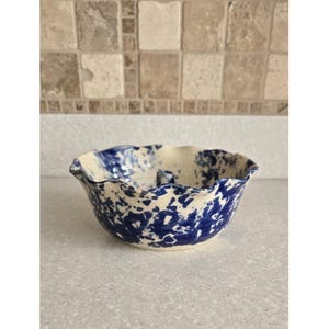 May include: A ceramic bowl with a cream-colored interior and a blue splatter design. The bowl has a wavy, uneven rim and sits on a light-colored surface. The background features a tile backsplash.