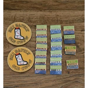 May include: A collection of ski patches and badges on a wooden surface. The patches are round and yellow with the text "SKI SAFELY OLD CROW" and a graphic of a ski boot. Rectangular badges display "BRONZE", "SILVER", and "NASTAR" with mountain names.