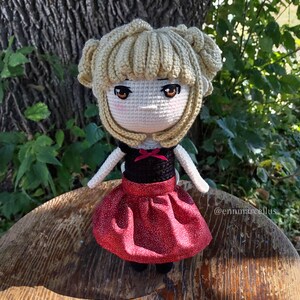 May include: Crocheted doll with blonde hair, wearing a black top with a red bow and a red skirt with white polka dots. The doll is sitting on a wooden surface.