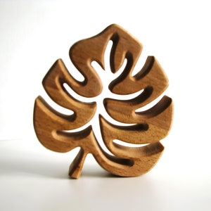 May include: A wooden leaf sculpture with a tropical monstera leaf design. The sculpture is carved from a light brown wood and features intricate cut-out details. The leaf stands upright, showcasing its natural wood grain.