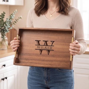May include: A rectangular wooden serving tray with a dark brown wood grain finish and gold-toned metal handles on the sides. The center of the tray features a laser-engraved design with a large letter W and the name WILKER printed in the middle.