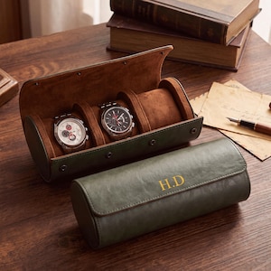 May include: A cylindrical olive green watch roll case with a snap closure and the gold monogram H.D. The interior is lined with brown suede and features three individual compartments, two of which contain silver-toned wristwatches with round dials.