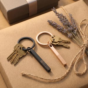 May include: Two keychains with keys, one black with "LOVE" engraved, and one rose gold with a date engraved. The keychains are on a brown surface with a gift box and lavender sprigs.