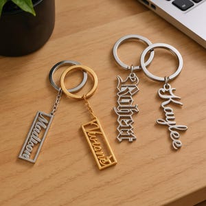 May include: Several personalised keyrings with names. The keyrings are silver and gold-coloured, featuring names such as "Madlond", "Viviane", "Robert", and "Kaylee" in stylish fonts. The keyrings are displayed on a wooden surface.