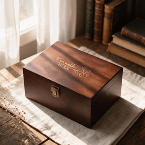 Puede incluir: Una caja rectangular de madera marrón oscuro con tapa abatible y un cierre de metal color latón. La tapa presenta un diseño floral grabado con el texto Kenneth y EST 2026. La caja tiene un acabado liso y un patrón de veta de madera visible.