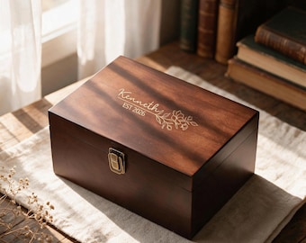 May include: A rectangular dark brown wooden box with a hinged lid and a brass-colored metal latch. The lid features an engraved floral design with the text Kenneth and EST 2026. The box has a smooth finish and a visible wood grain pattern throughout.