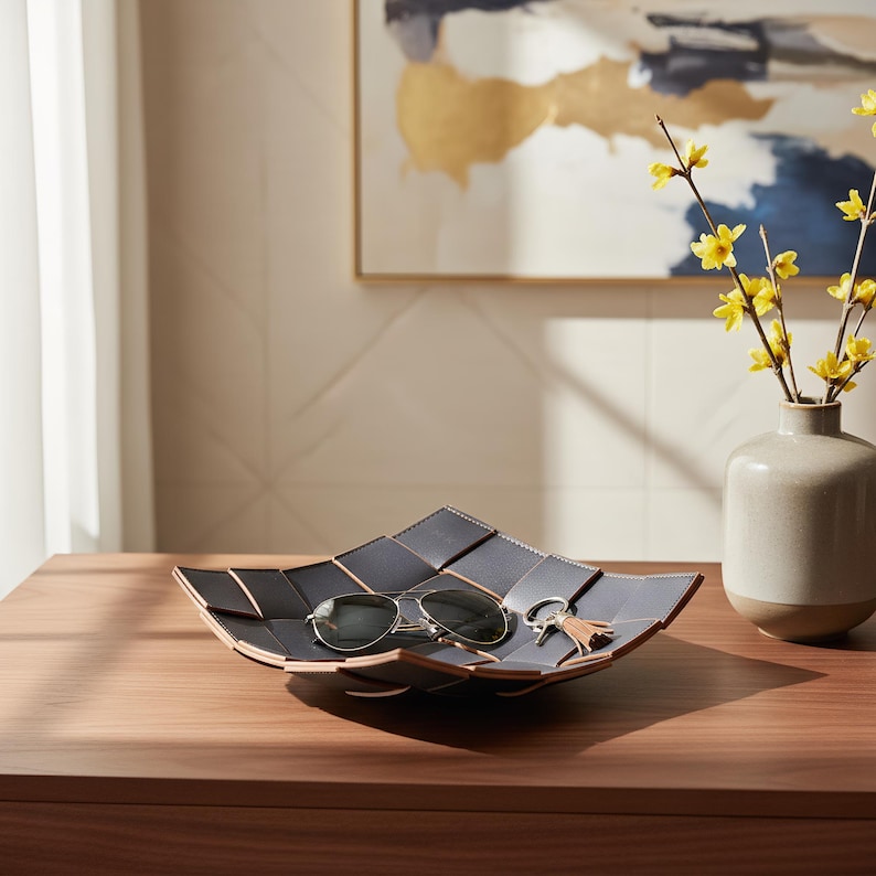 May include: A square, dark gray woven leather tray with visible stitching along the edges. The tray features a shallow, multi-paneled design and contains a pair of metal-framed sunglasses and a small keychain with a light brown leather tassel.