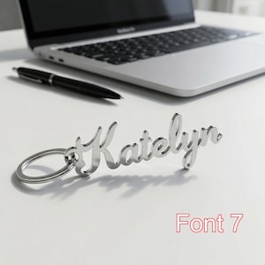 May include: A silver-colored keychain with the name "Katelyn" in a cursive font. The keychain is on a white surface, with a laptop and pen in the background. The text "Font 7" is in red.