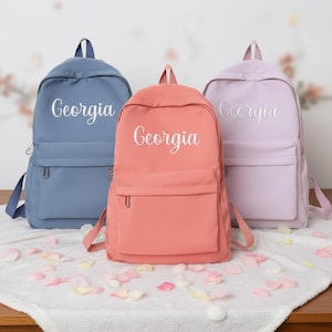 Puede incluir: Tres mochilas en tonos azul, coral y lavanda. Cada mochila tiene el nombre "Georgia" bordado en blanco. Las mochilas están sobre una superficie blanca con pétalos de rosa esparcidos.