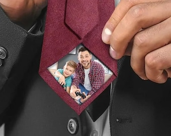 May include: Burgundy tie with a diamond-shaped inset featuring a photo of a man and a child playing a video game. The tie is held by a person wearing a black suit.