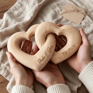 Puede incluir: Dos corazones de madera de color claro entrelazados con un acabado suave y veteado natural. El corazón izquierdo está grabado con el texto Phoebe & Spike, y el derecho con el texto Est. 2026 en una fuente cursiva de color oscuro.