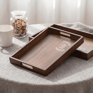 May include: A rectangular dark brown wooden serving tray with cutout handles on the short sides. The center of the tray features a white engraved design consisting of the letters MK surrounded by a laurel wreath leaf pattern.