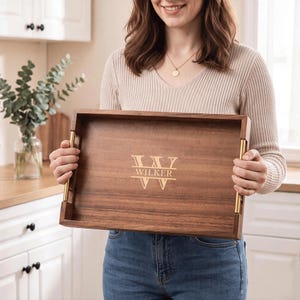 May include: A rectangular wooden serving tray with a dark brown finish and gold-toned metal handles on the sides. The centre of the tray features a laser-engraved design with a large letter W and the name WILKER printed horizontally across it.