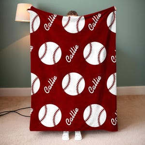 May include: A maroon blanket with a repeating pattern of white baseballs with red stitching. The name "Callie" is printed in white script between the baseballs. The blanket is held up by a person.