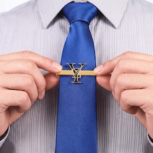 May include: A solid blue necktie featuring a gold-toned metal tie clip. The tie clip is adorned with a prominent, stylized black and gold letter Y emblem centered on the horizontal bar, secured against a light grey pinstriped dress shirt.