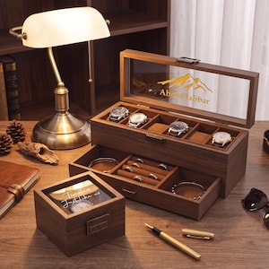 May include: A dark wood watch and jewelry storage box with a glass lid featuring a mountain graphic and the name Abdul Jabbar. The box includes a pull-out drawer with velvet-lined compartments for rings and bracelets, alongside a smaller matching box.