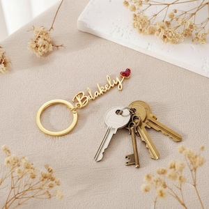 May include: A gold-colored keychain with the name "Blakely" in cursive script, topped with a red heart-shaped charm. Several keys of varying sizes and colors are next to the keychain, suggesting a personalized accessory for organizing keys.