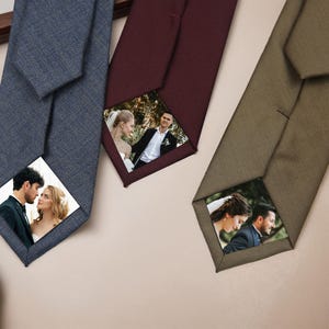 Op de afbeelding: Drie stropdassen in blauwe, bordeauxrode en olijfgroene tinten, elk met een gepersonaliseerde foto aan de punt. De stropdassen zijn gerangschikt op een lichtbeige oppervlak, met een textuurstof. De foto's lijken van stellen te zijn.
