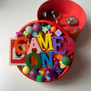 May include: A red round box with a colorful game themed design. The box is open and the inside is filled with colorful beads, dice, and game pieces. The lid of the box has the words "GAME ON" spelled out in colorful letters.