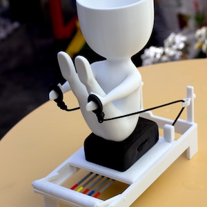 May include: A white, stylized figure with a hollow head sits on a black rectangular block atop a white frame structure. The figure holds black cords attached to the frame, which features four thin rods colored gray, yellow, blue, and red.