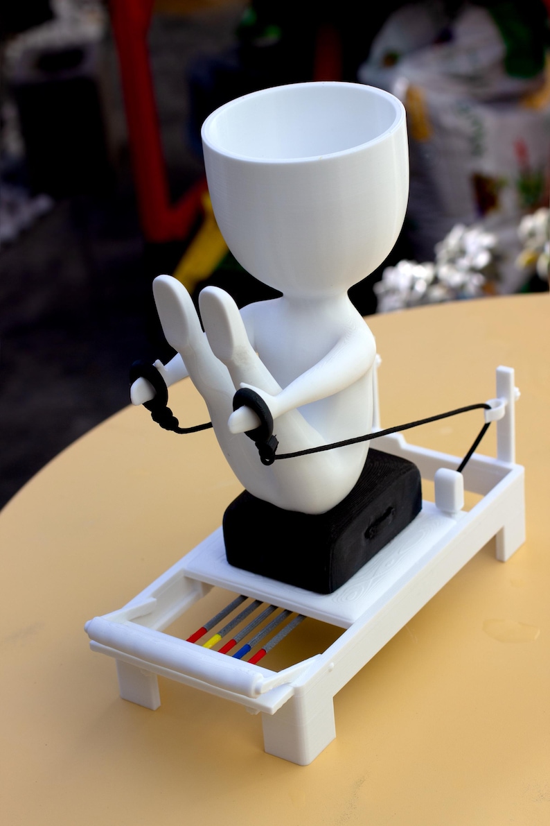 May include: A white, stylized figure with a hollow head sits on a black rectangular block atop a white frame structure. The figure holds black cords attached to the frame, which features four thin rods colored gray, yellow, blue, and red.
