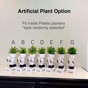 May include: Seven white planters labeled A through G, each featuring a figure performing Pilates exercises with black resistance bands. Each planter holds a small green artificial plant and sits on a white base. Text reads Artificial Plant Option.
