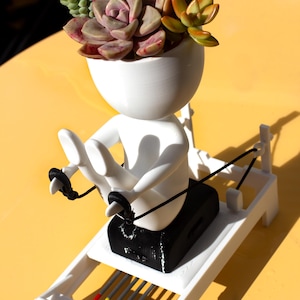 May include: A white planter shaped like a stylized figure using a rowing machine, with a head filled with a variety of green, red, and orange succulents. The rowing machine base is white with black cords and four small colored rods in red, yellow, and blue.