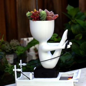 May include: A white planter shaped like a stylized human figure sitting on a rowing machine. The figure has a round head containing a cluster of small, multi-colored succulent plants. The rowing machine base is white with black straps and accents.